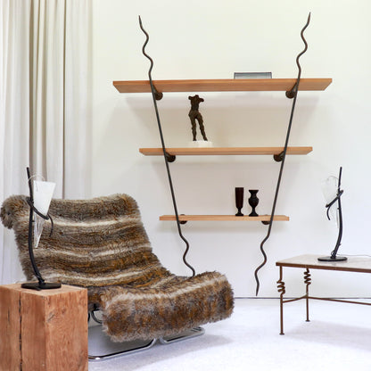 Shelving unit in a room setting with chair, lamp and table.