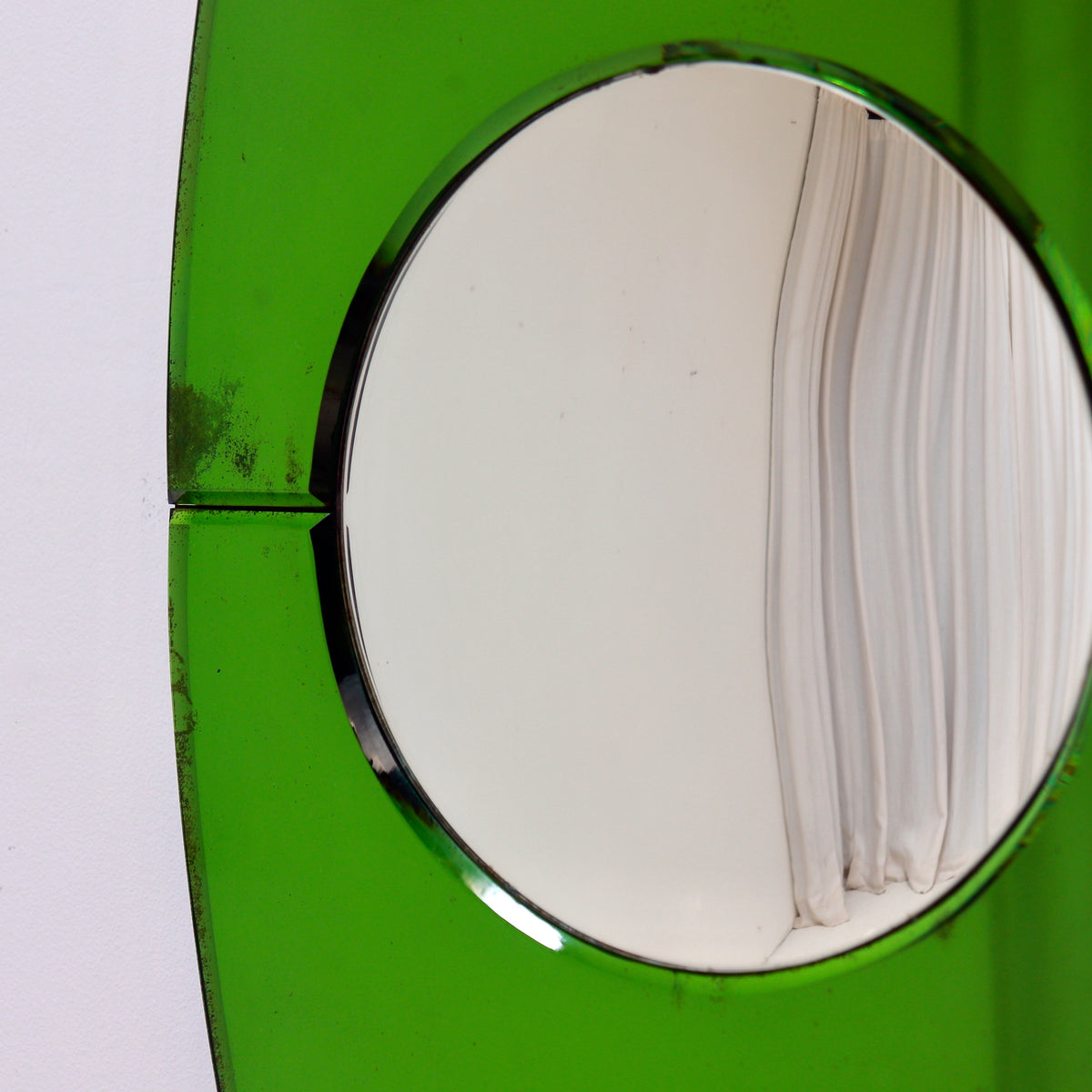 A close-up of a bright green oval mirror, reflecting a white curtain on one side and a plain light background on the other.