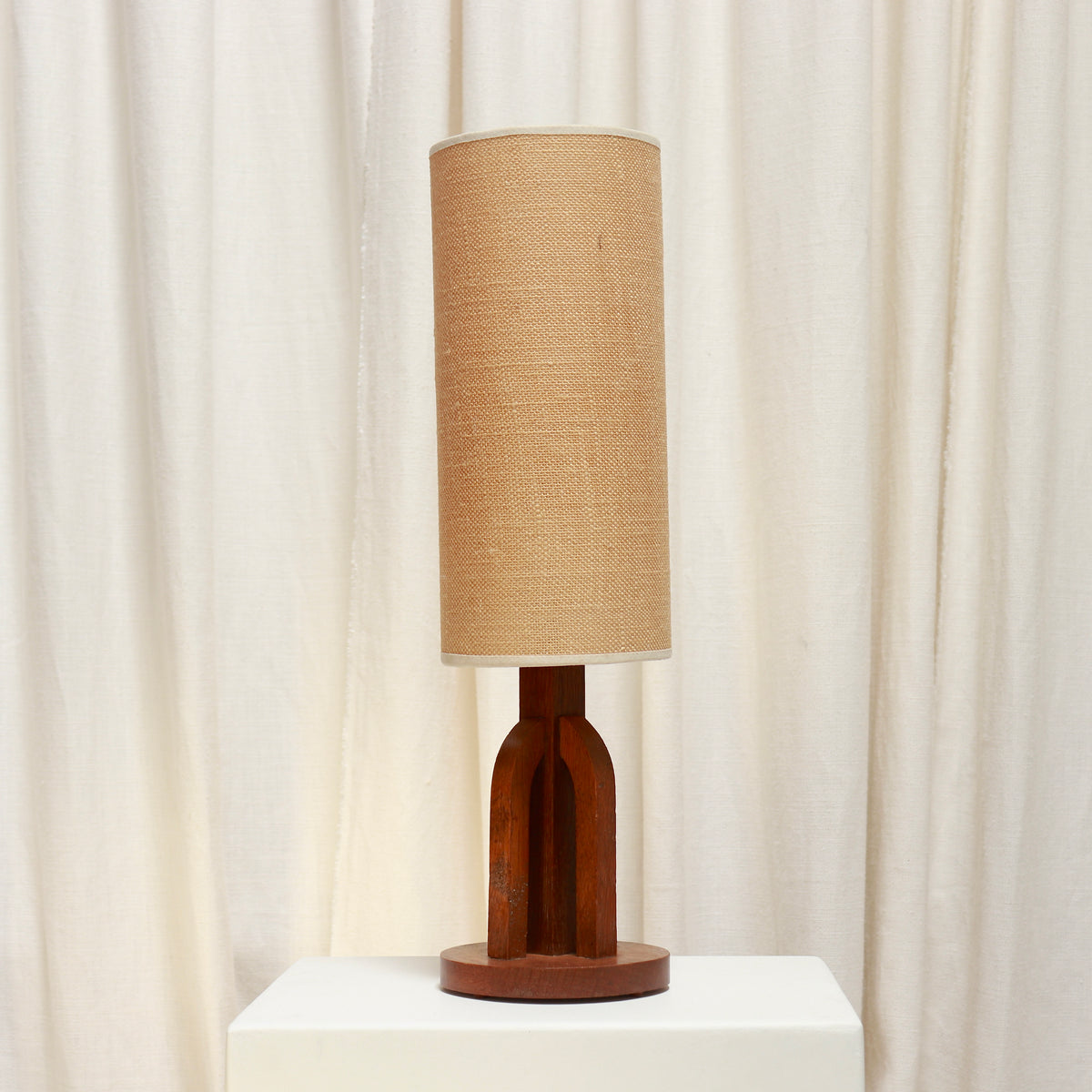 A table lamp with a cylindrical woven beige fabric shade and a sculptural dark brown wooden base, displayed on a white pedestal in front of draped cream curtains.
