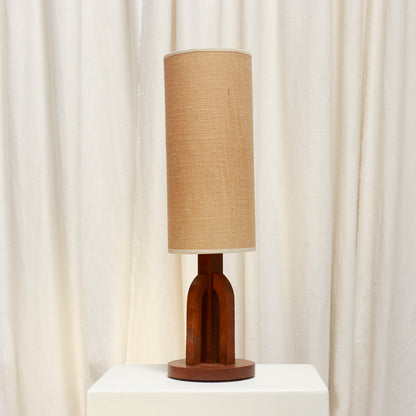 A table lamp with a cylindrical woven beige fabric shade and a sculptural dark brown wooden base, displayed on a white pedestal in front of draped cream curtains.