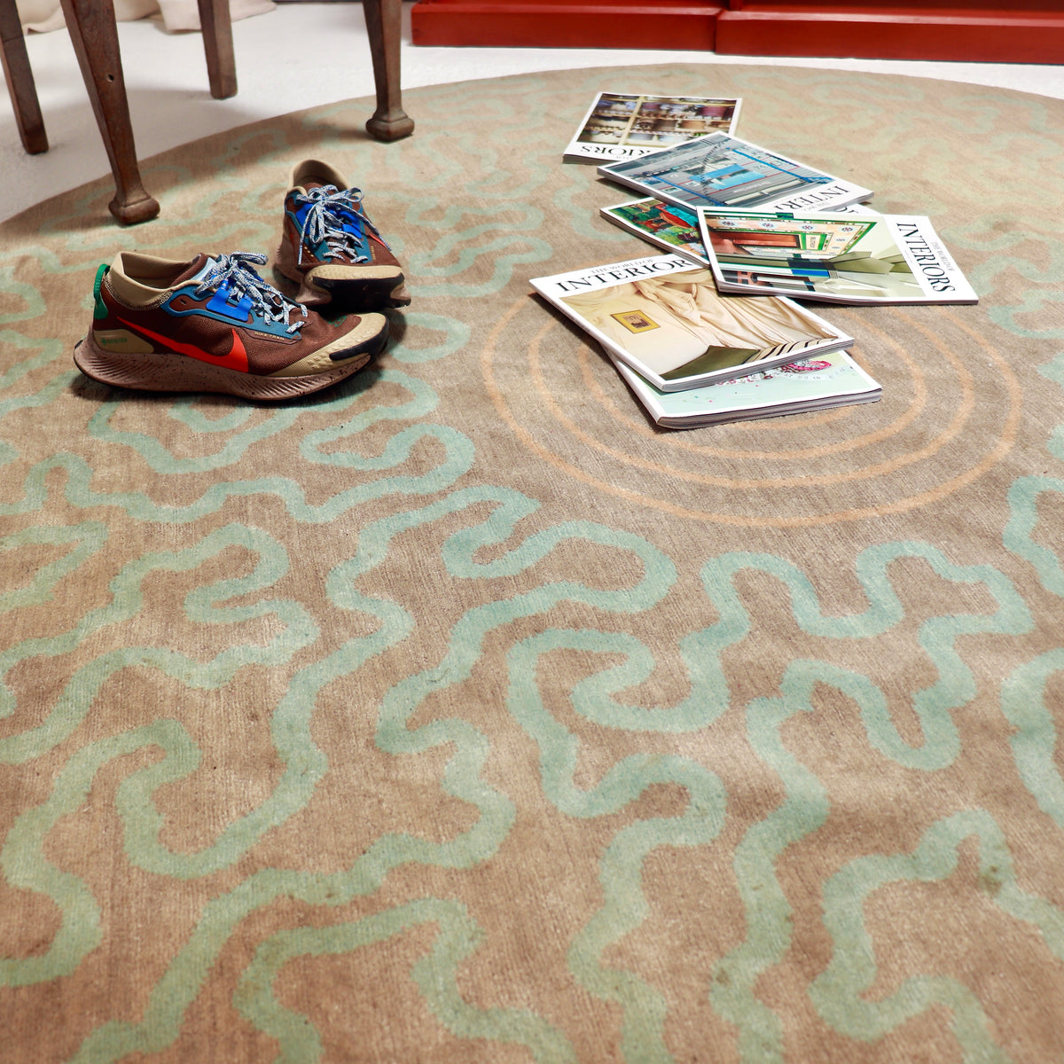 Circular wool rug styled with magazines and trainers, showing aqua squiggles and peach concentric rings on a taupe ground.