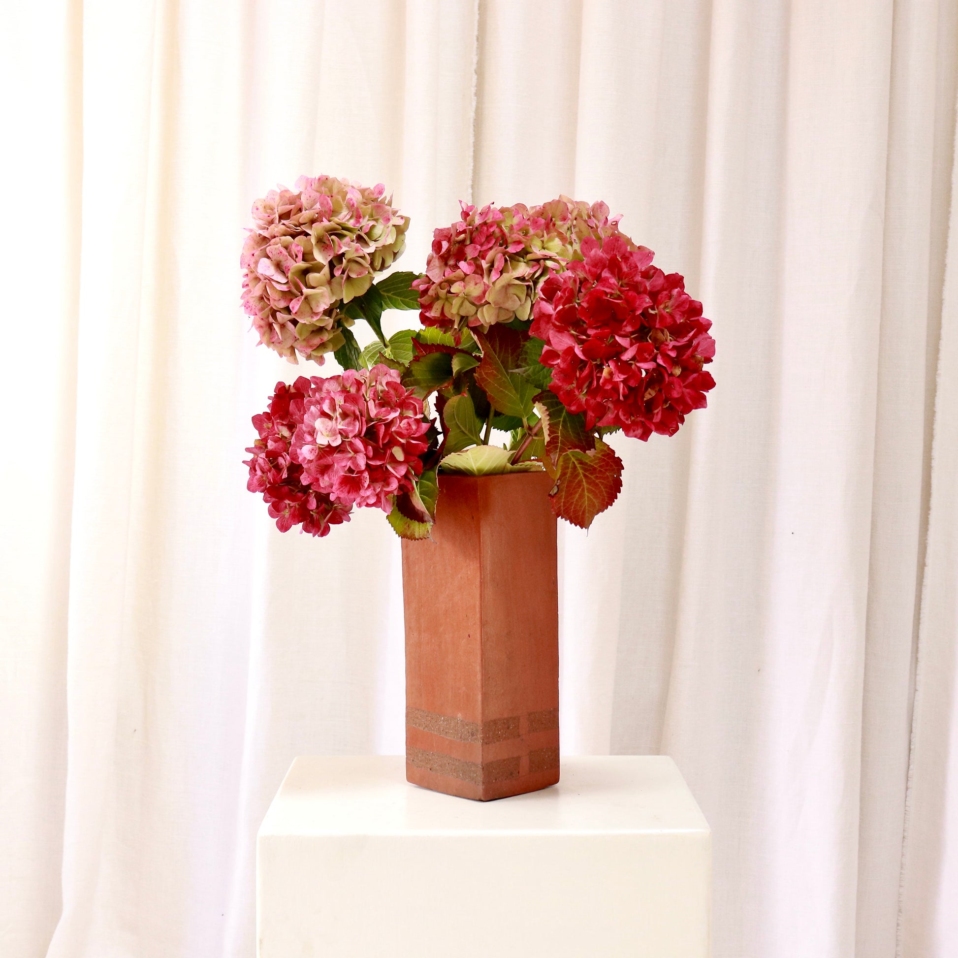Tall hand-built terracotta slab vase with banded base holding hydrangeas on a plinth.