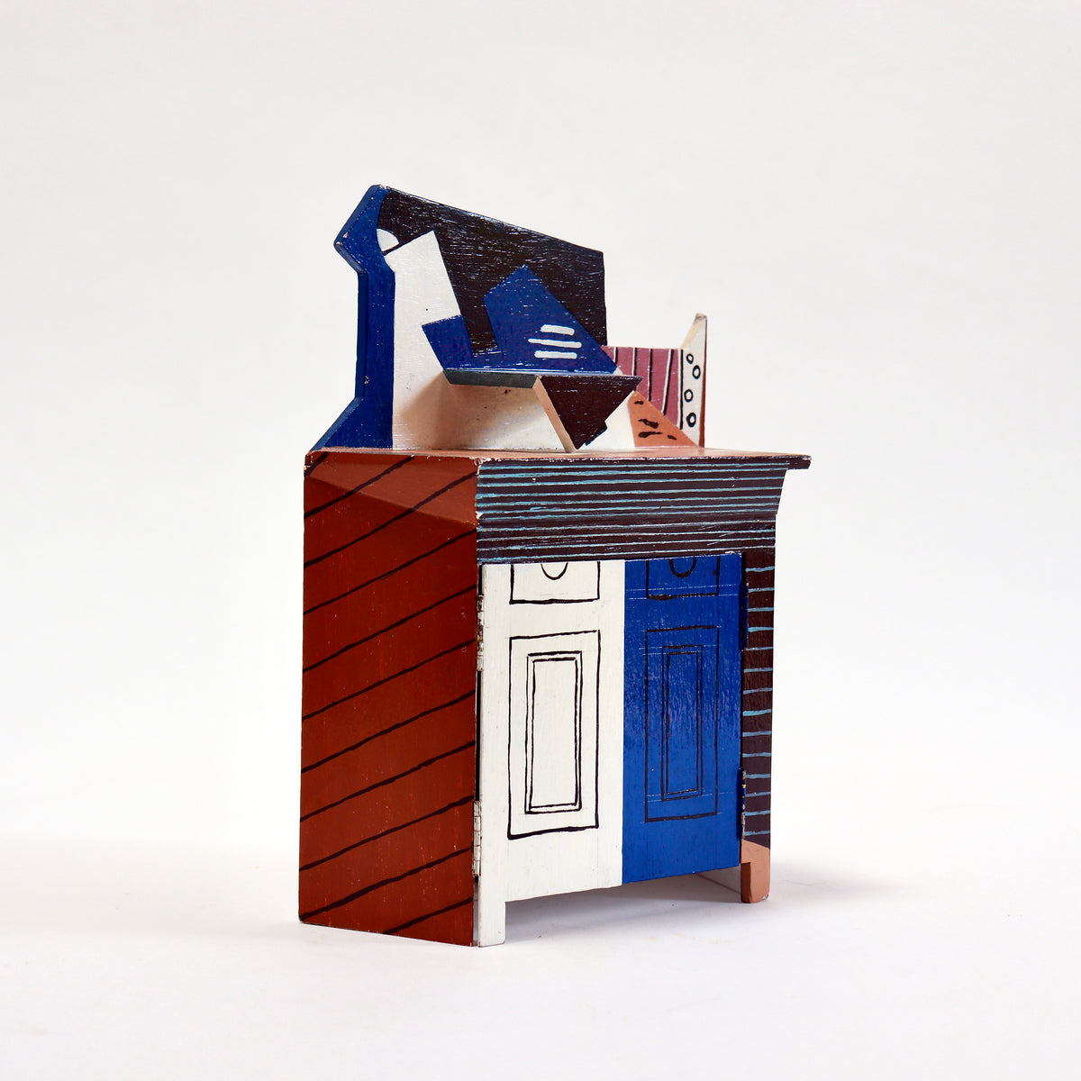 A handcrafted miniature house model made from layered cardboard, featuring painted details in blue, red-brown, white, and black, set against a plain white studio background.