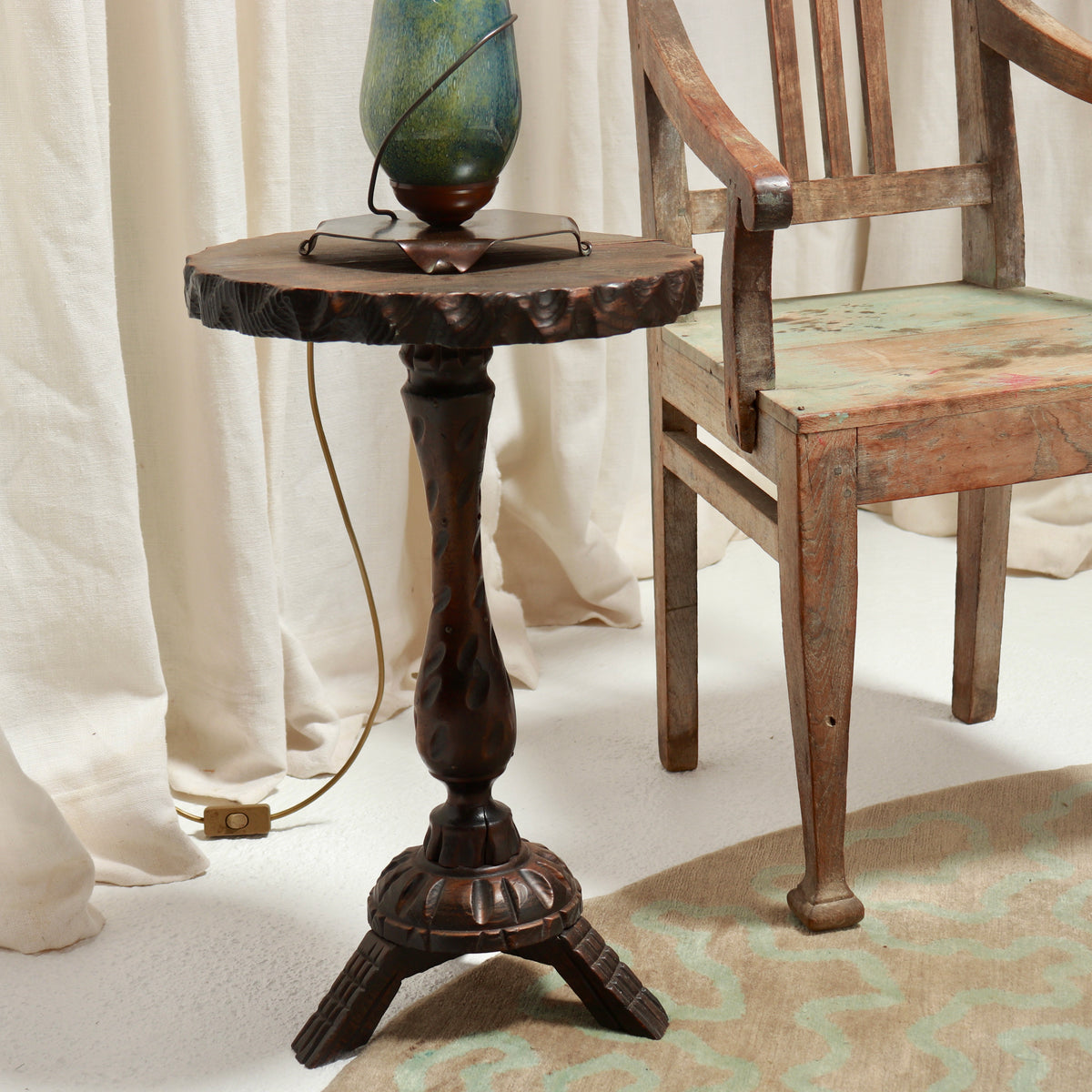 A rustic dark brown wooden side table with a carved pedestal base and a rough-sawn top, styled next to a wooden chair with draped cream fabric in the background.