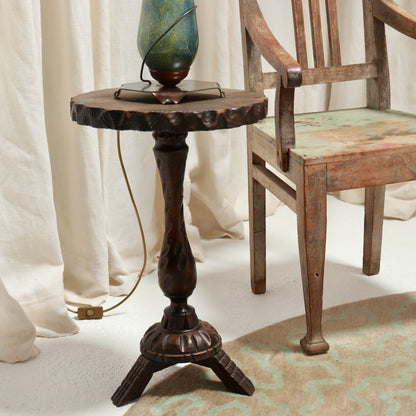 A rustic dark brown wooden side table with a carved pedestal base and a rough-sawn top, styled next to a wooden chair with draped cream fabric in the background.