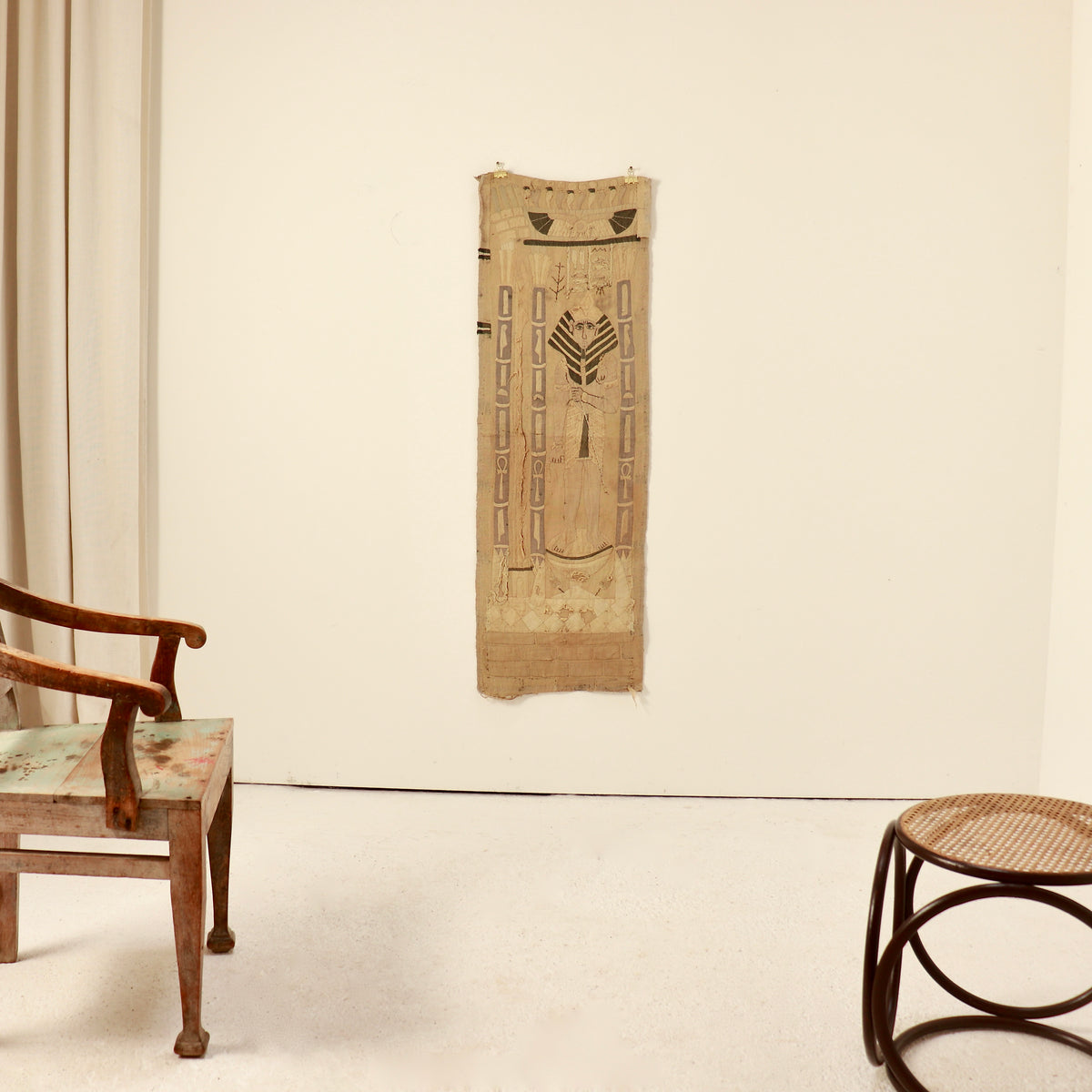 A vertical woven textile wall hanging with geometric and tribal-style patterns in natural tan and brown, displayed on a plain off-white wall in a room with a wooden chair and stool.