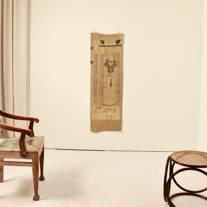 A vertical woven textile wall hanging with geometric and tribal-style patterns in natural tan and brown, displayed on a plain off-white wall in a room with a wooden chair and stool.