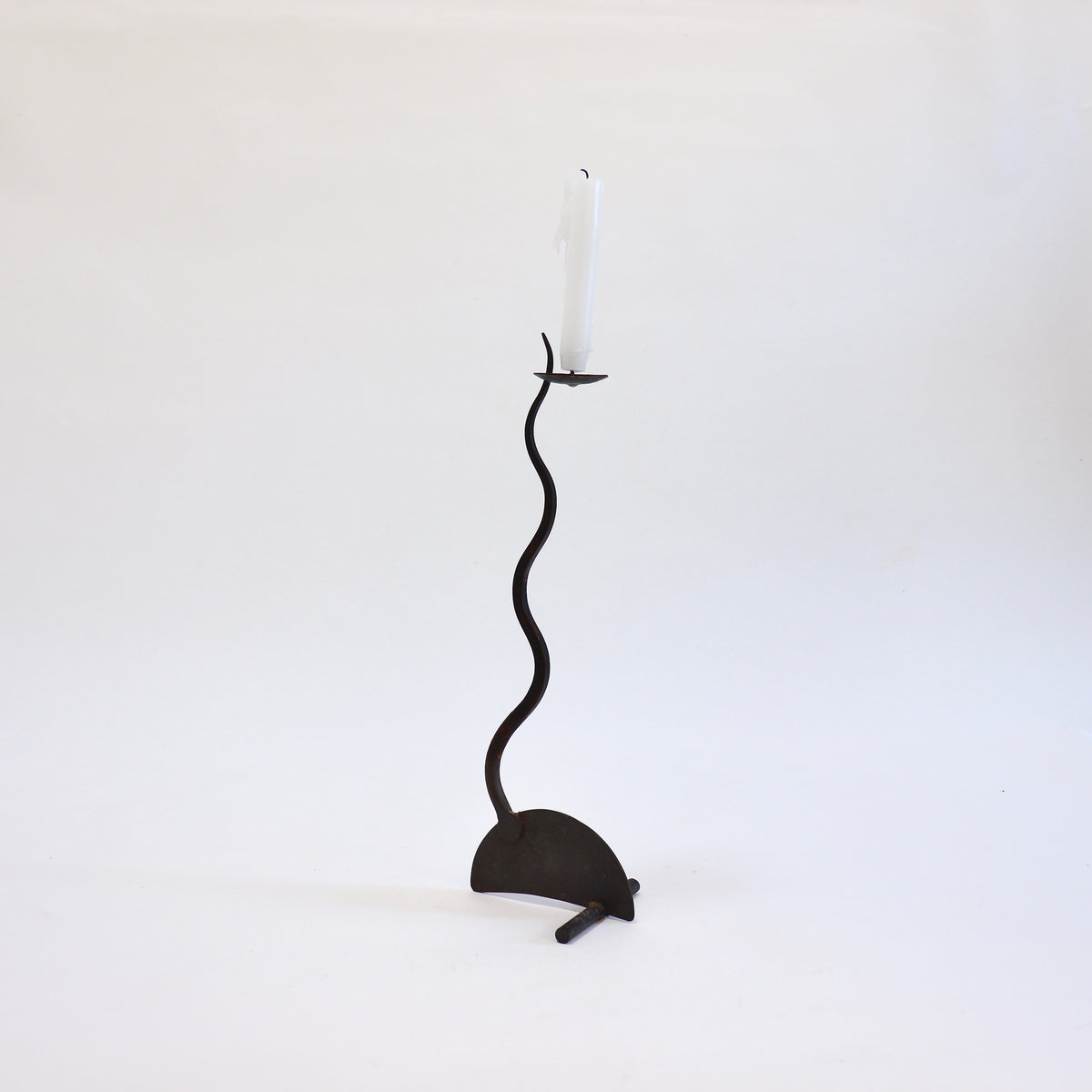 Hand-forged iron candlestick with a snaking stem, semicircular base and white taper candle.