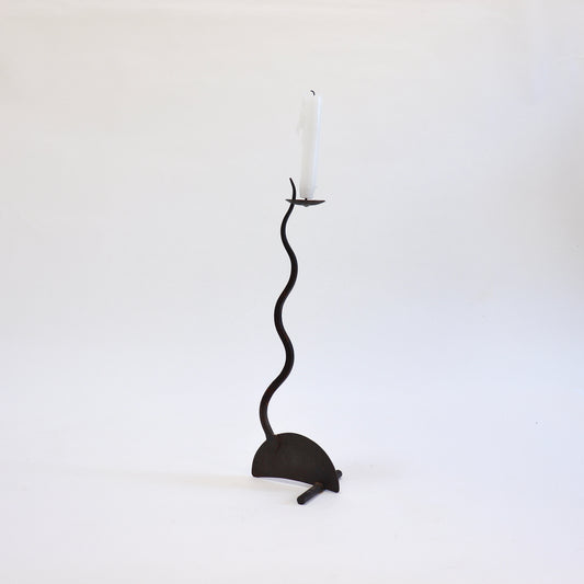 Hand-forged iron candlestick with a snaking stem, semicircular base and white taper candle.