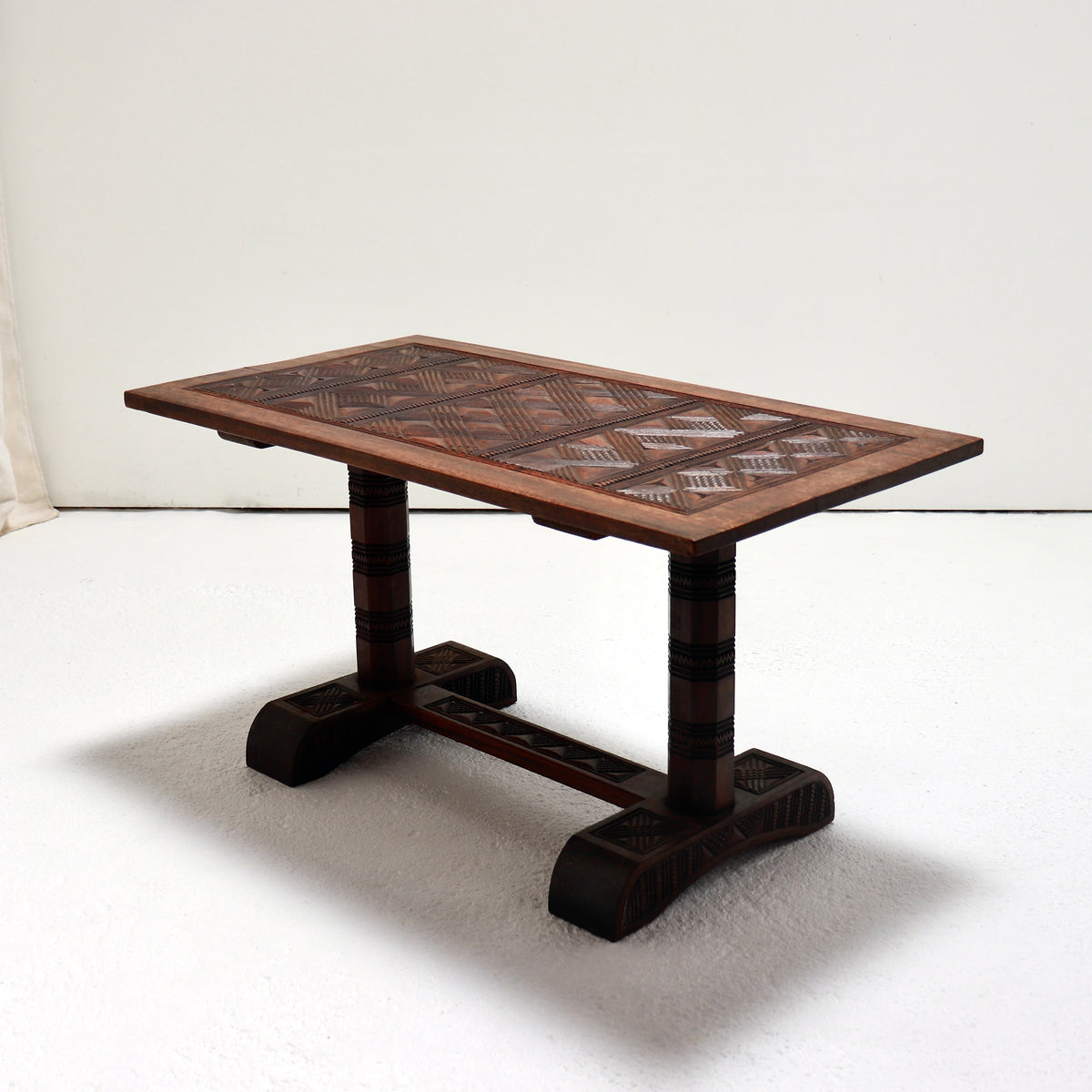 Three-quarter view of a Nigerian hardwood coffee table with a geometric chip-carved top and carved trestle base.