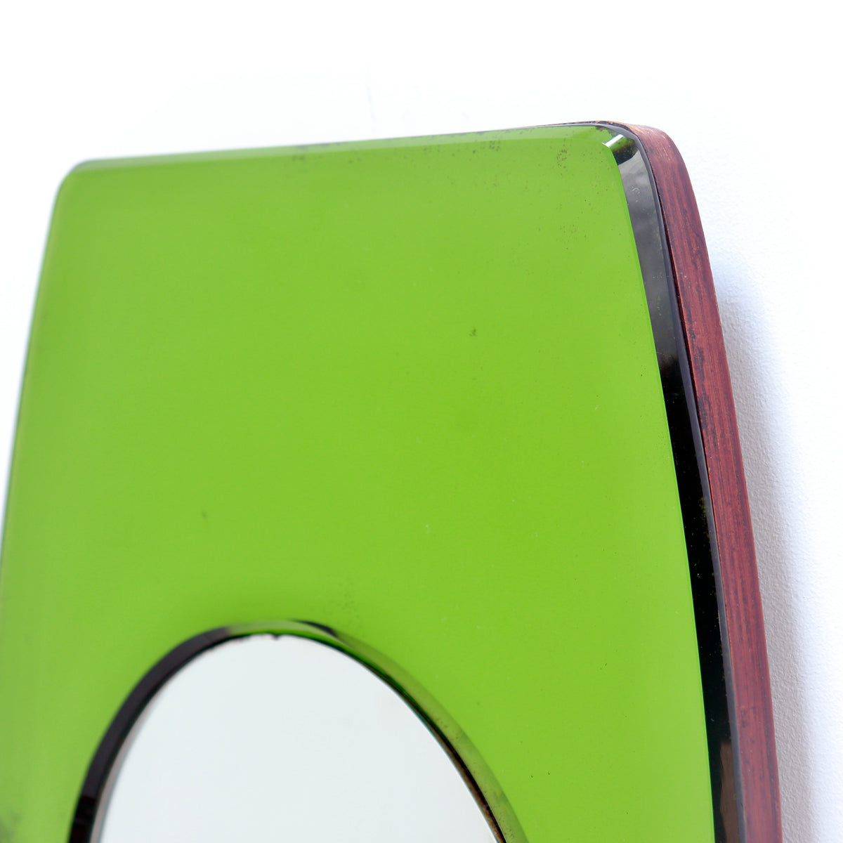 Close-up of an art deco wall mirror with a bright lime green glass panel and a dark reddish-brown wood frame, showing a small portion of the circular mirror reflection.