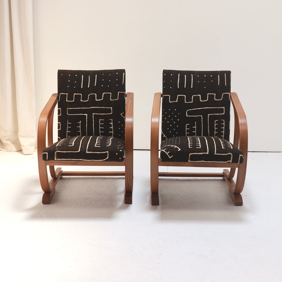 Front view of a pair of 1930s Heal’s armchairs with warm bentwood frames and dark mud cloth upholstery patterned with pale geometric motifs.