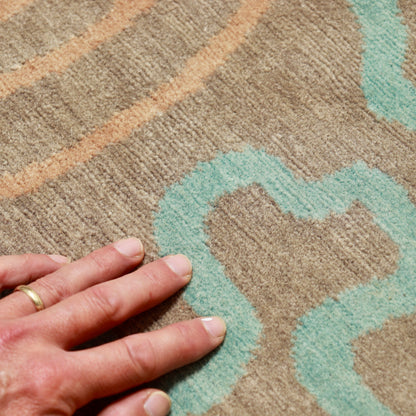 Hand beside the rug’s short wool pile, showing its texture and aqua and peach pattern.