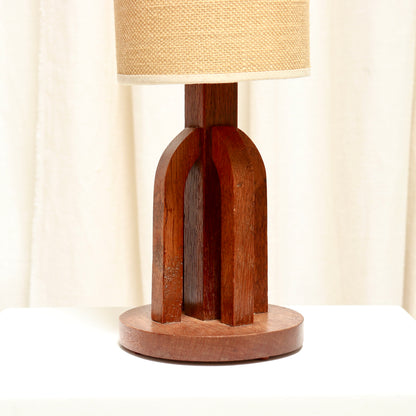 An art deco oak table lamp with a warm reddish-brown carved base and a textured beige fabric drum shade, set against a soft cream curtain background.