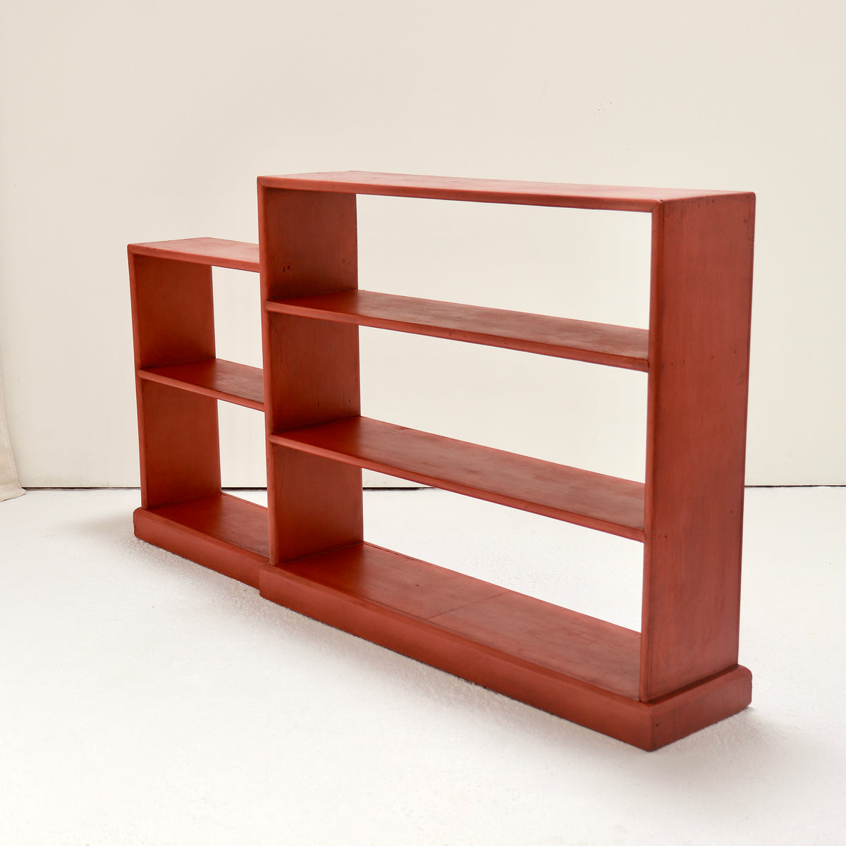 A mid-century modern wooden bookcase with a warm reddish-brown finish, featuring three tiers of open shelving on the right and two cubby sections on the left, set against a plain off-white background.
