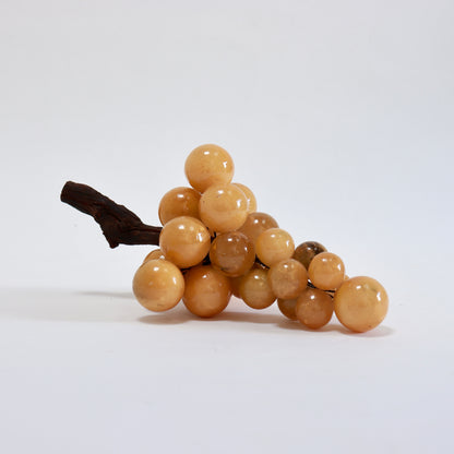 Large alabaster grape bunch resting horizontally, with golden stone fruit and a gnarled wooden stem extending left.