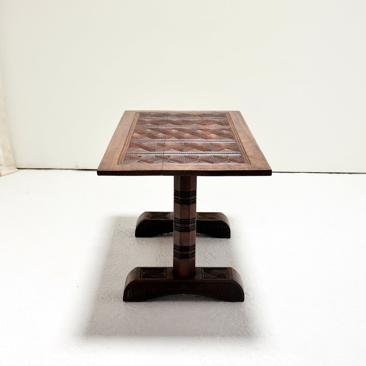 End view of the hardwood coffee table showing the patterned top, faceted support and broad carved foot.
