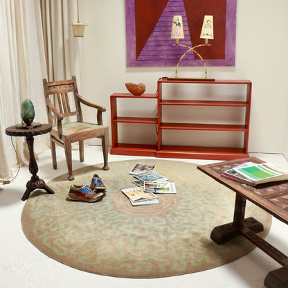 A round area rug with a beige and sage green wavy pattern, styled in a mid-century modern living room setting with a wooden chair, shelving, and other decor.