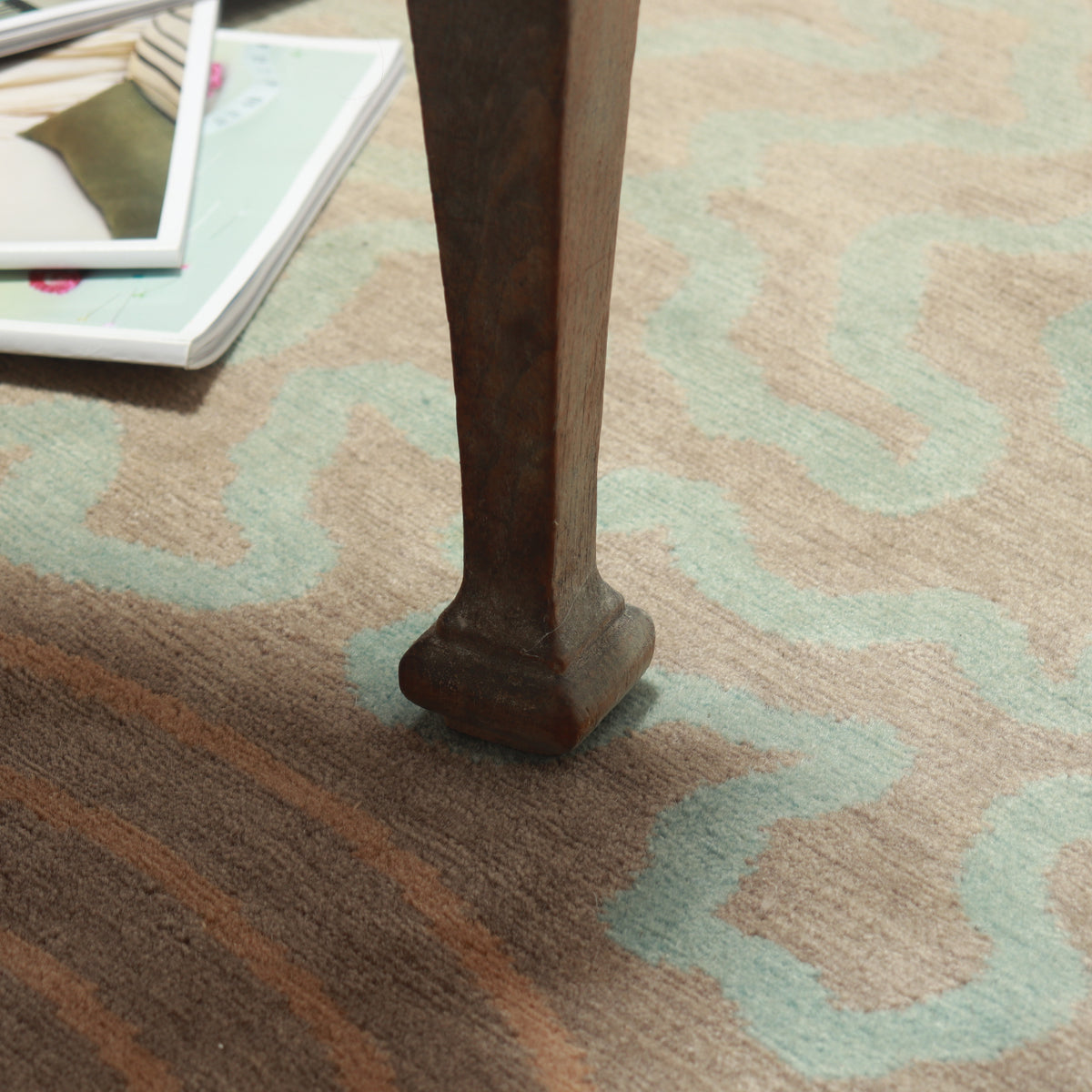 Detail of the patterned wool rug beneath a wooden chair leg, with magazines nearby.