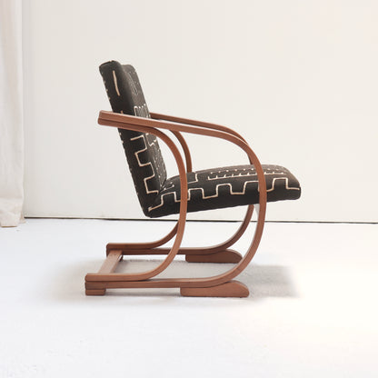 Angled view of a Heal’s Art Deco armchair with curved wooden arms and geometric antique mud cloth upholstery.