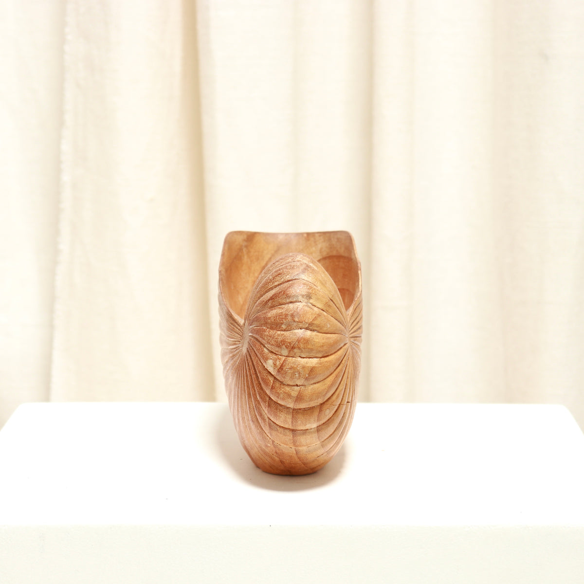A hand-carved wooden cup with a textured, ribbed exterior, displayed on a white surface against a soft beige curtain background.