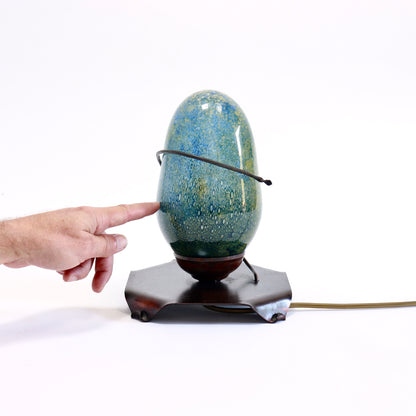 A vintage-style table lamp with a glossy speckled blue and green ceramic egg-shaped shade, supported by a dark brown metal stand. A person's hand points to the lamp, and a power cord is visible on a white background.