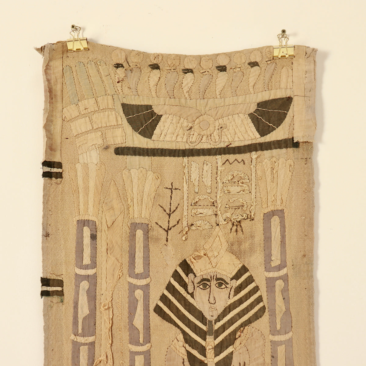 An ancient Egyptian-style textile tapestry or wall hanging, featuring hieroglyphic motifs, a pharaoh figure, and a winged sun emblem, suspended by two gold-colored metal clips against a plain off-white wall.
