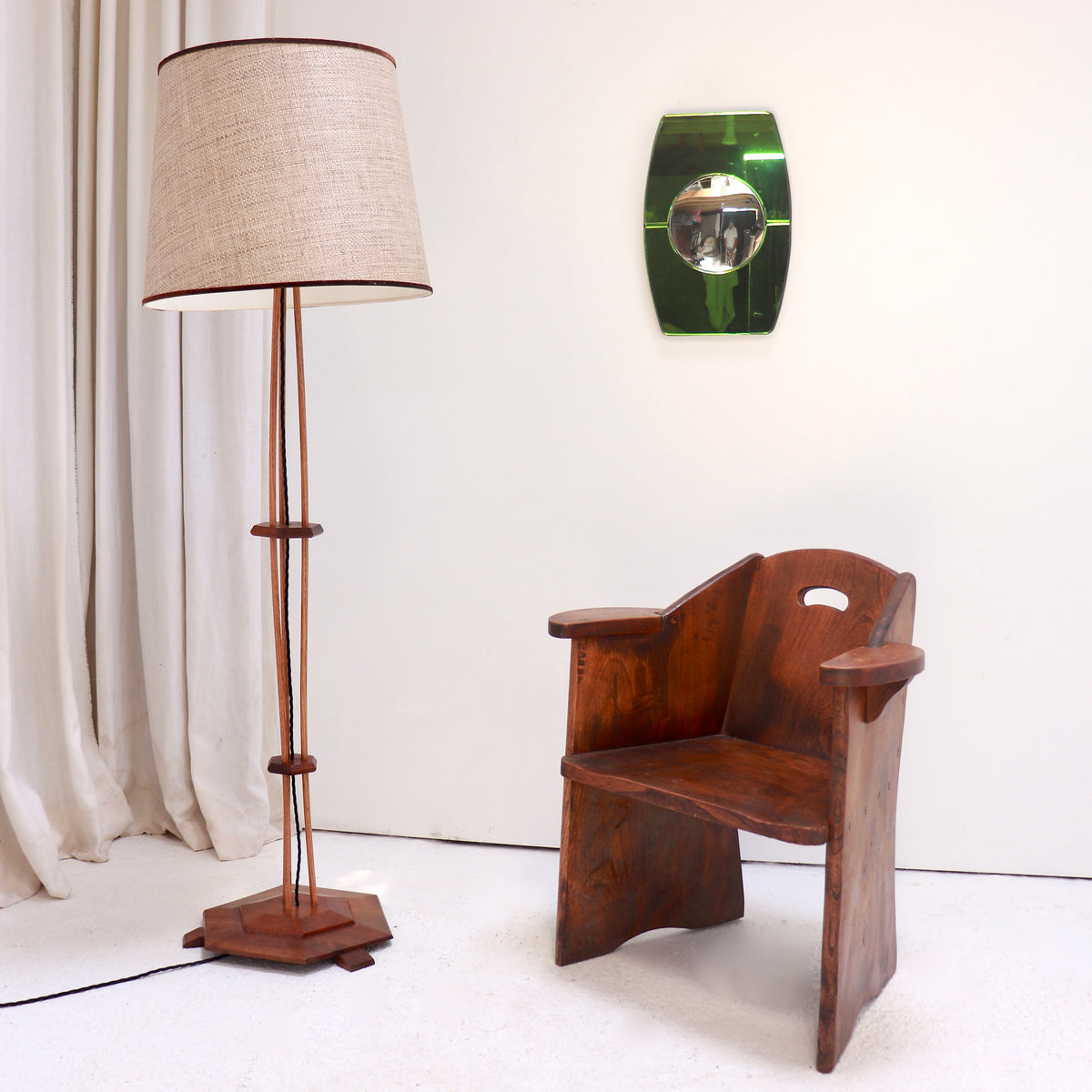 An early 20th century wooden armchair with a curved back and armrests, shown next to a tall floor lamp with a beige linen shade and a green wall mirror on a white wall.