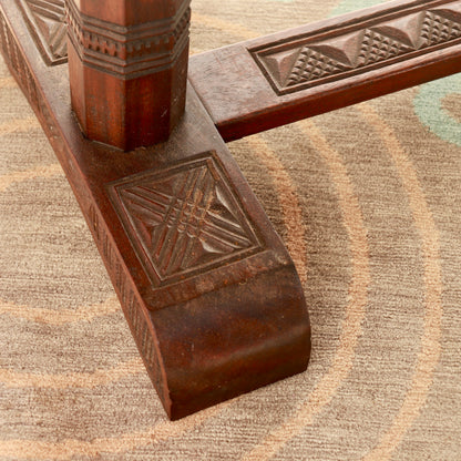Close-up of a carved dark brown wooden table base with geometric patterns, resting on a textured beige and brown patterned area rug.