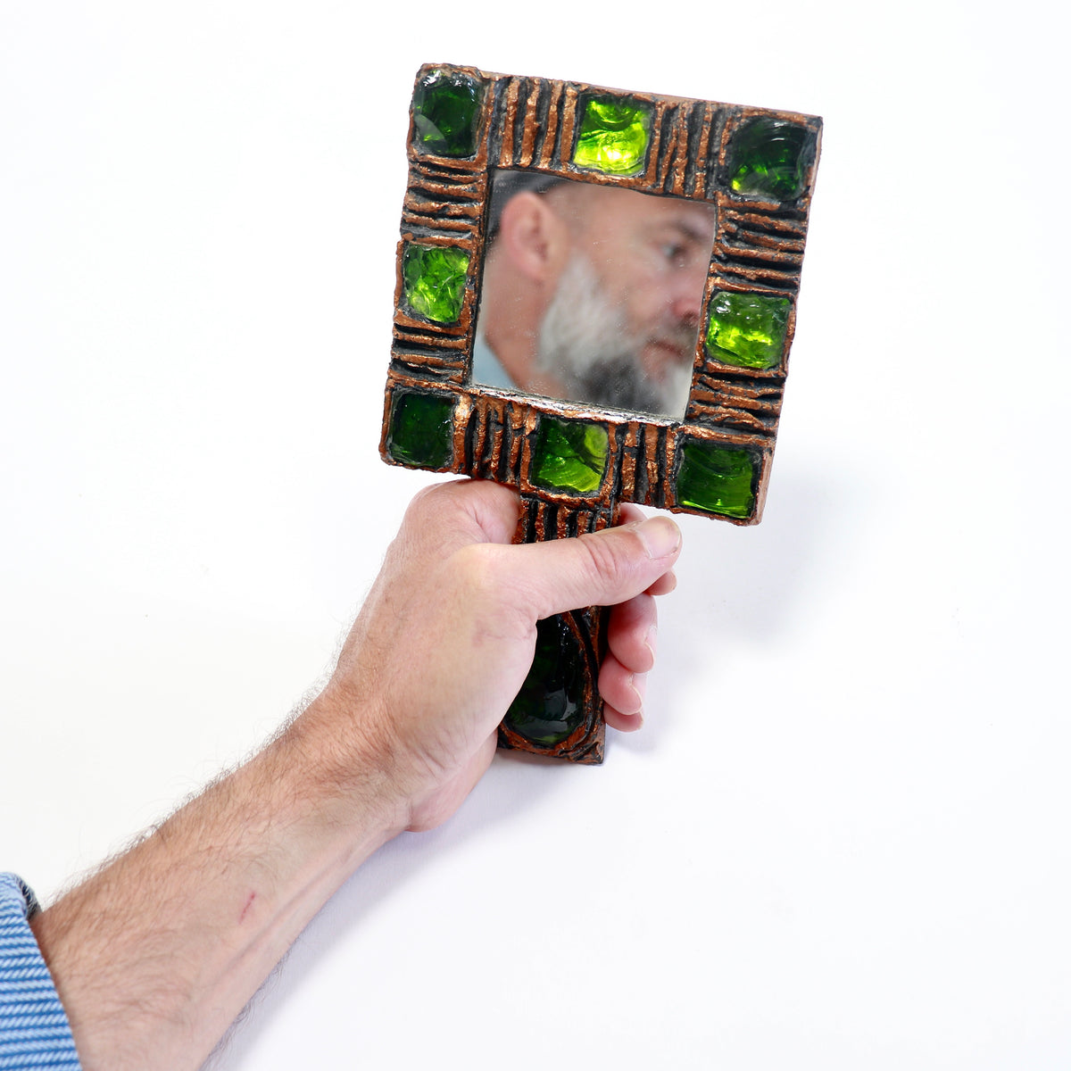 A hand holding a square decorative hand mirror with a rustic brown frame accented by green glass tiles, showing a reflection of a bearded man against a white background.
