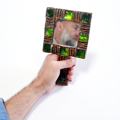 A hand holding a square decorative hand mirror with a rustic brown frame accented by green glass tiles, showing a reflection of a bearded man against a white background.