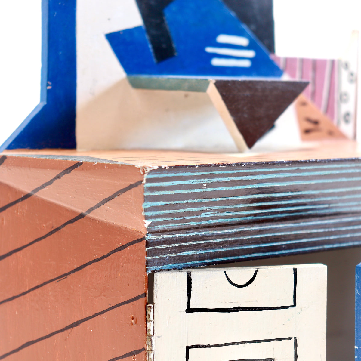 Close-up of a handcrafted wooden box or miniature structure with geometric cutout details, painted in terracotta, blue, white, and black.