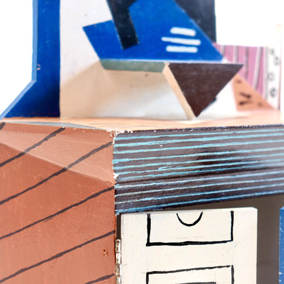 Close-up of a handcrafted wooden box or miniature structure with geometric cutout details, painted in terracotta, blue, white, and black.