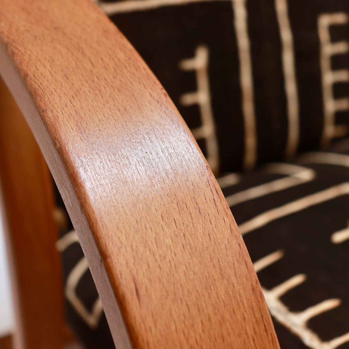 Detail of a restored Heal’s armchair’s curved wooden arm, showing the exposed grain beside dark patterned mud cloth upholstery.