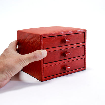A small red leather jewelry box with three drawers and matching drawer pulls, held by a person's hand against a plain white background.