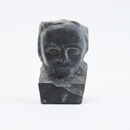 Front view of the slate portrait bust showing almond-shaped eyes, a closed mouth and integral stone base.