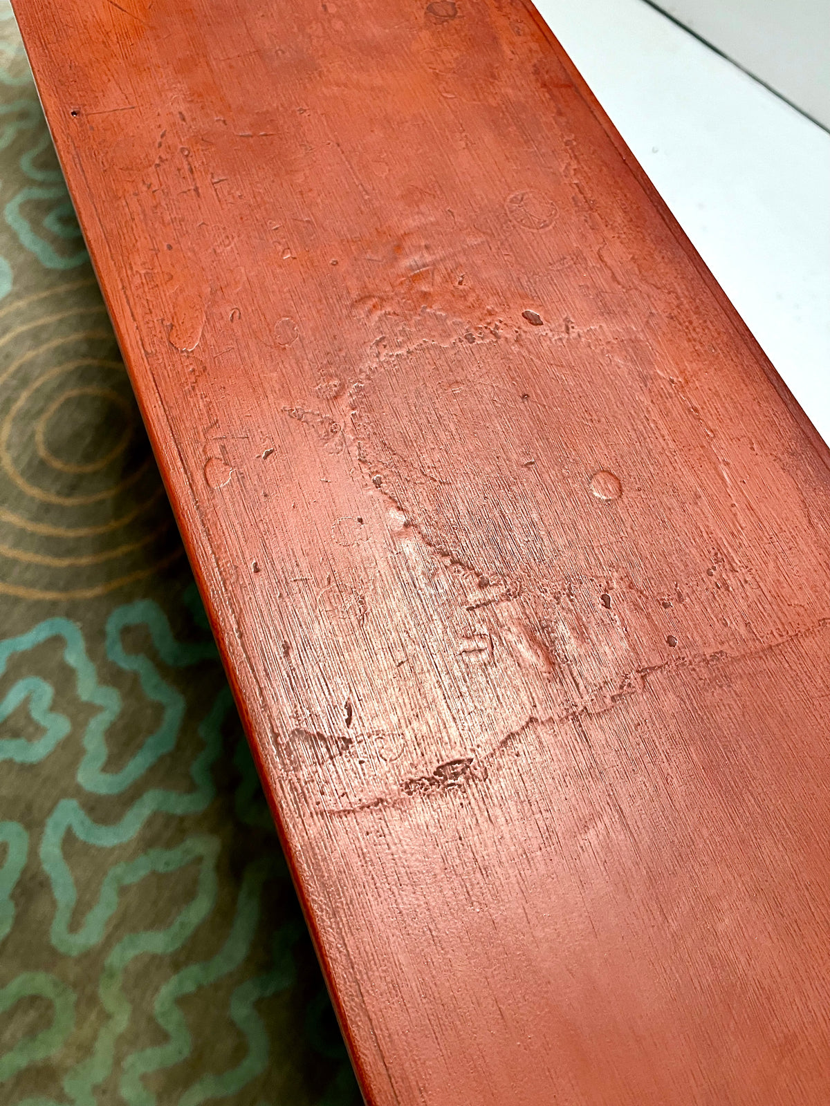 A close-up of a reddish-brown wooden board or furniture piece with visible wear, scratches, and a chipped area, placed over a patterned green rug.