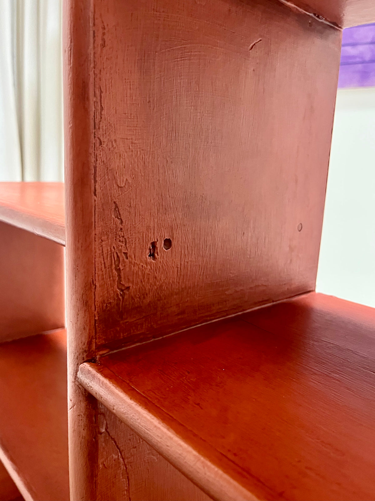 Close-up of a reddish-brown wooden shelf or bookcase corner with visible wear, scratches, and small holes on the painted wood surface.