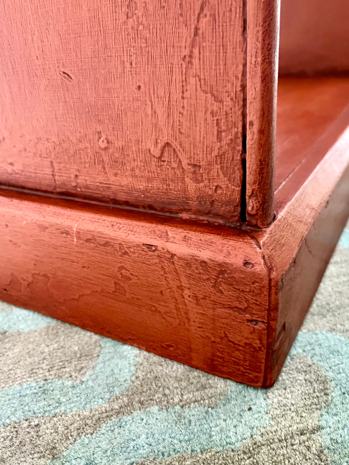 Close-up of a reddish-brown wooden furniture corner showing the distressed wood grain and a seam on a patterned rug.